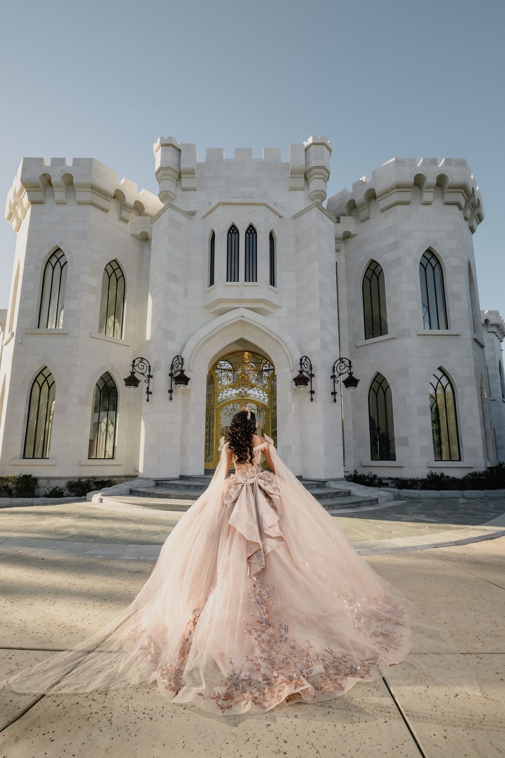 A woman in a flowing pink ballgown stands before a white castle-like building with Gothic arched windows and gold doors.