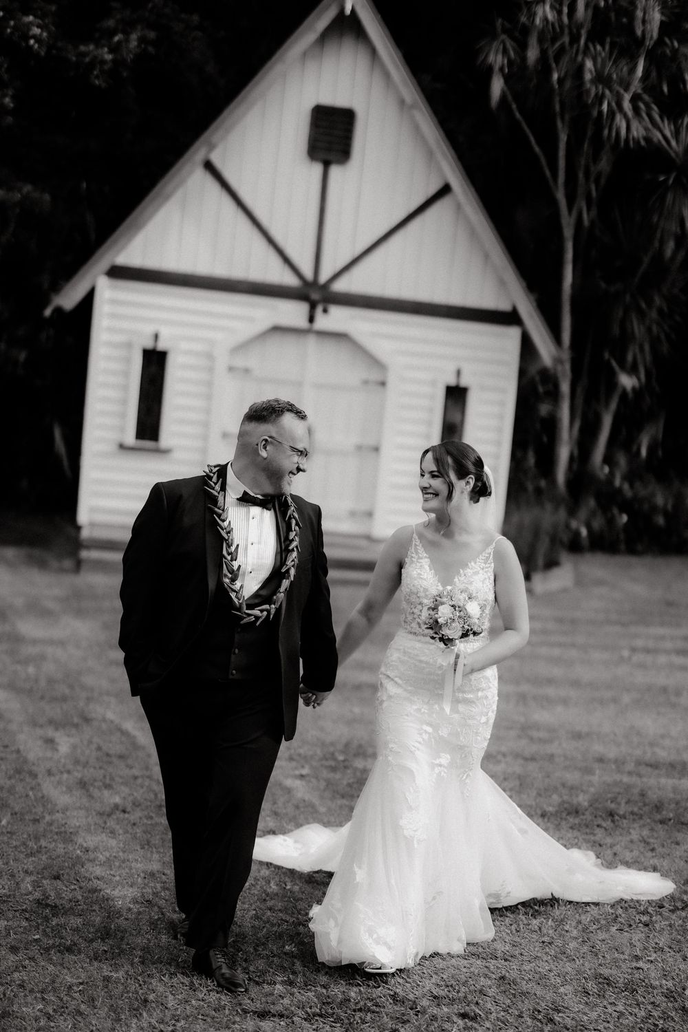 Black and white photo of a bride and groom walking hand in hand in front of a white chapel.