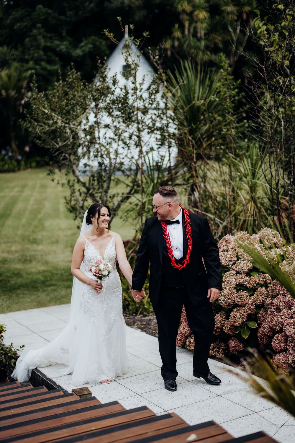 Bride and groom walk hand in hand through a lush garden venue, groom wearing a red lei over his black tuxedo.