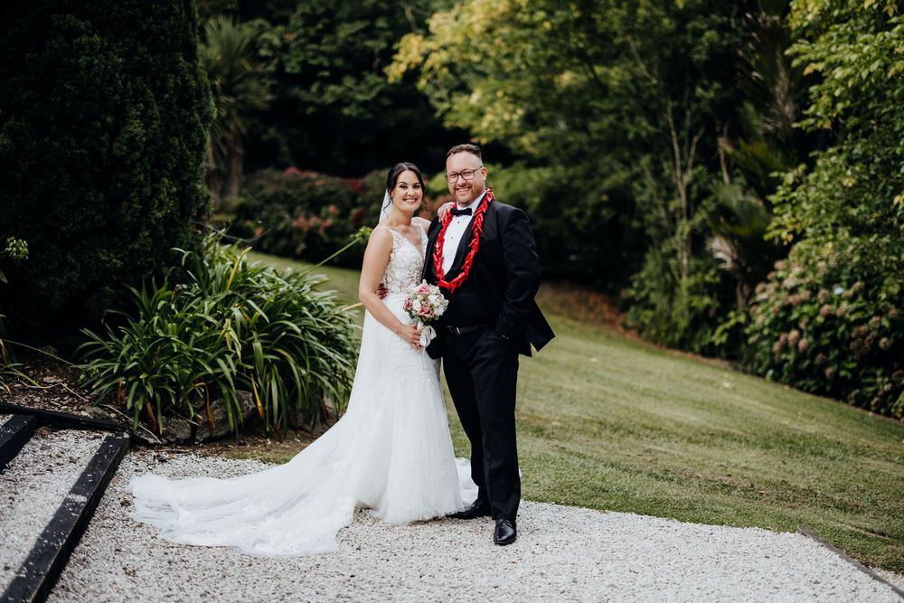 Bride in elegant white gown poses with groom in black tuxedo on a gravel path in a beautiful garden.