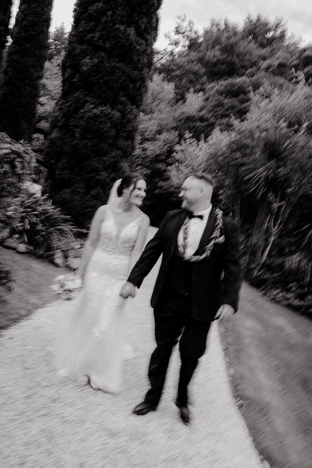 Black and white photo of a bride and groom walking hand in hand along a garden path lined with tall cypress trees.