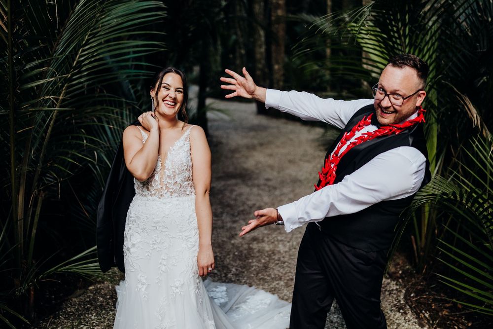 Bride in white gown laughs joyfully as groom in vest and floral lei gestures dramatically outdoors.