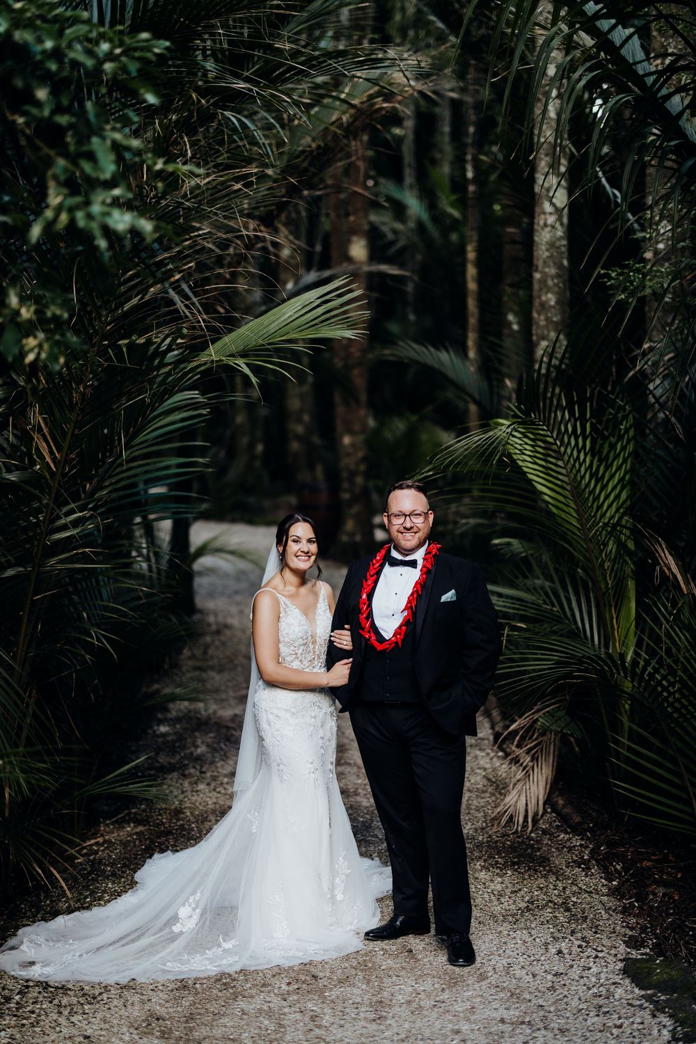 Bride and groom pose on a palm-lined gravel path, she in a lace gown, he in a tuxedo with red lei.