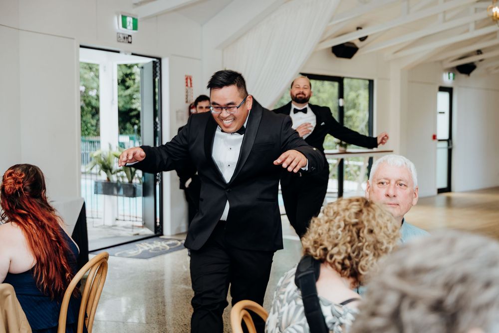 Groom in black tuxedo makes grand entrance at wedding reception, dancing through doorway with guests seated nearby.