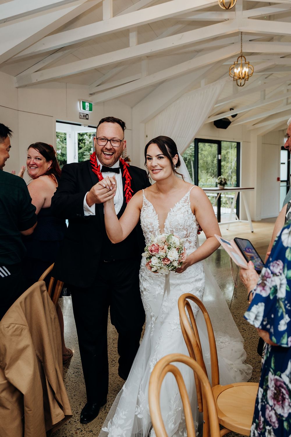 Newlyweds walk joyfully down the aisle, bride holding a bouquet, groom wearing a red lei in bright venue.