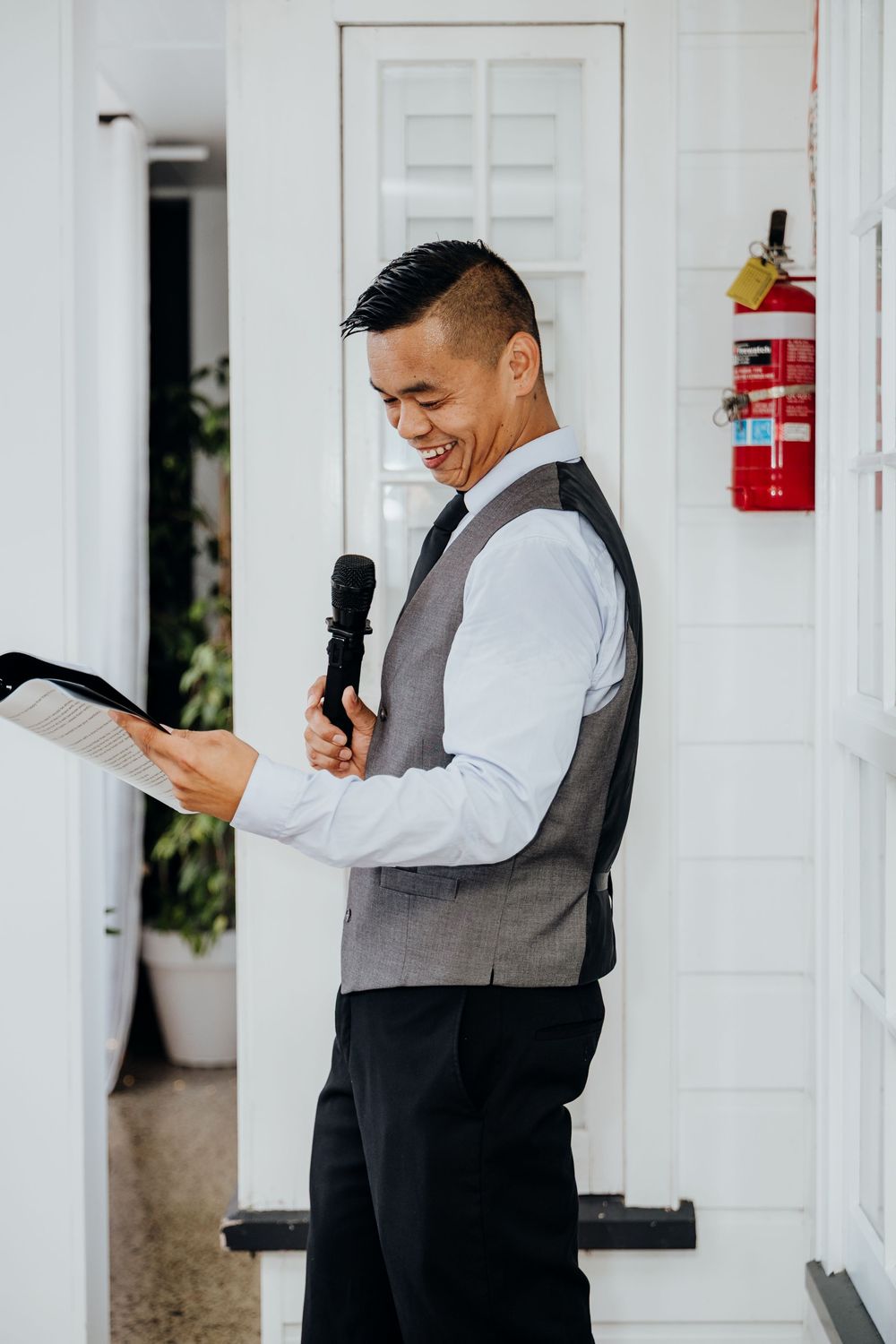 Smiling man in vest giving a speech at a wedding reception while holding a microphone and papers.