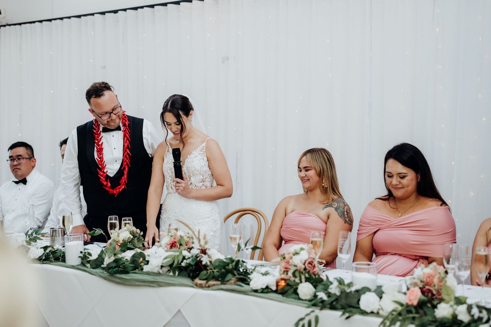 Bride and groom stand at wedding reception head table with bridesmaids in pink dresses, floral garland centerpiece.