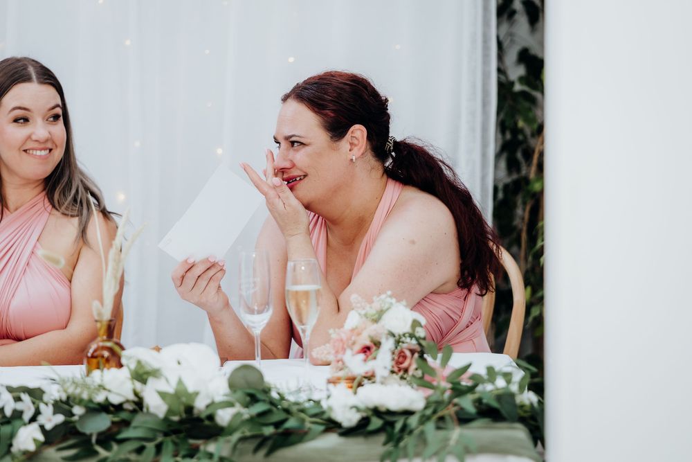 Bridesmaid in pink dress wipes tears of joy during emotional wedding speech at reception table with flowers.