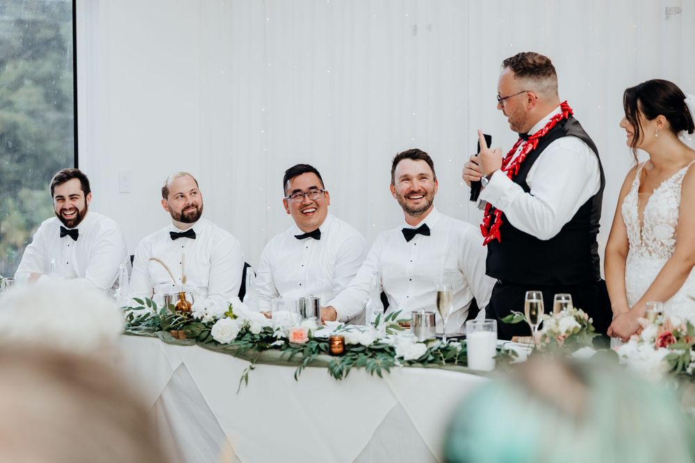 Groom and groomsmen laughing at wedding reception head table while officiant gives speech to bride and groom.