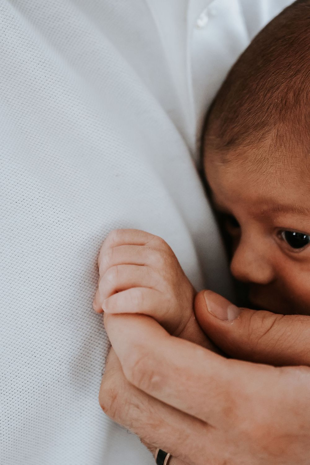 A newborn baby nestled against an adult in white, tiny hand grasping a finger in a tender moment.