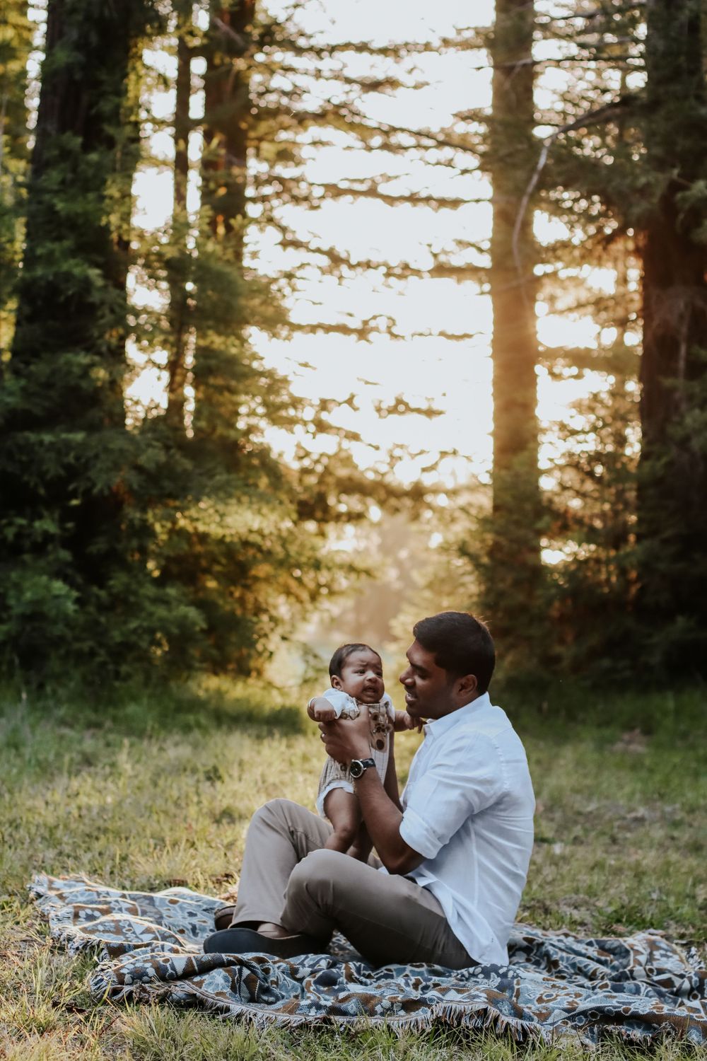 Father sits on blanket in sunlit forest holding smiling baby, sharing a joyful outdoor moment together.