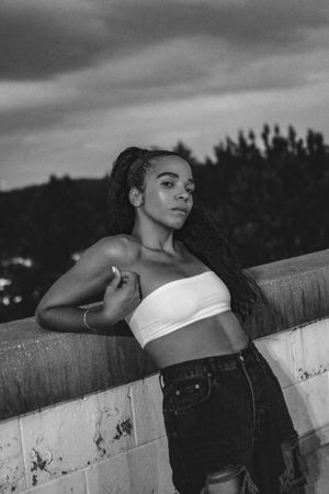 Young Black woman in white crop top and jeans leaning on wall, moody black and white portrait.