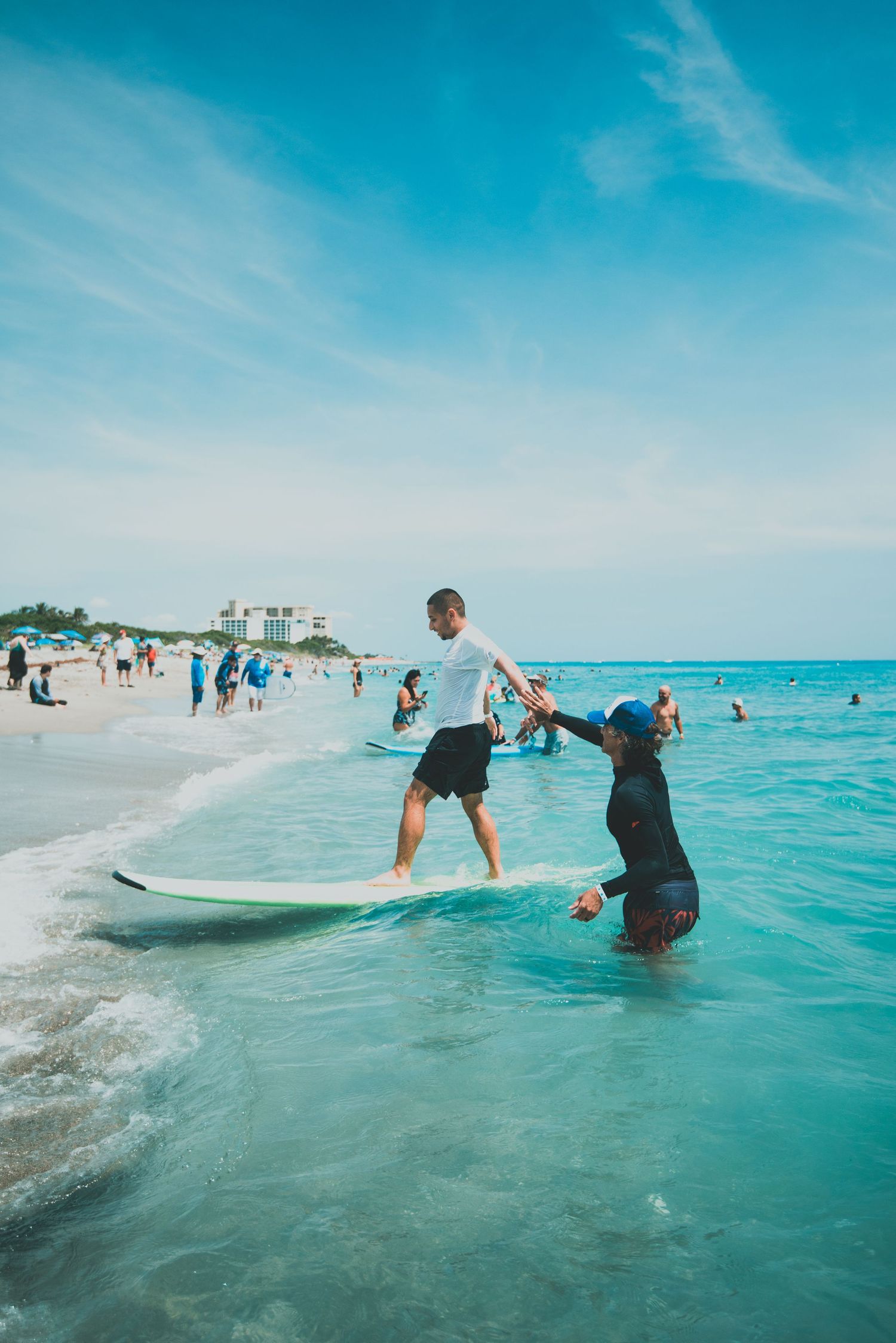 Man balancing on surfboard while instructor holds his hand in shallow turquoise ocean water on a sunny beach day.