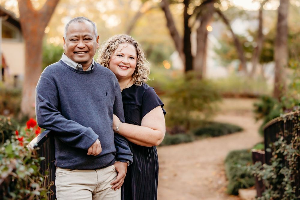 A smiling couple poses together on a garden path surrounded by trees and red flowers in warm evening light.