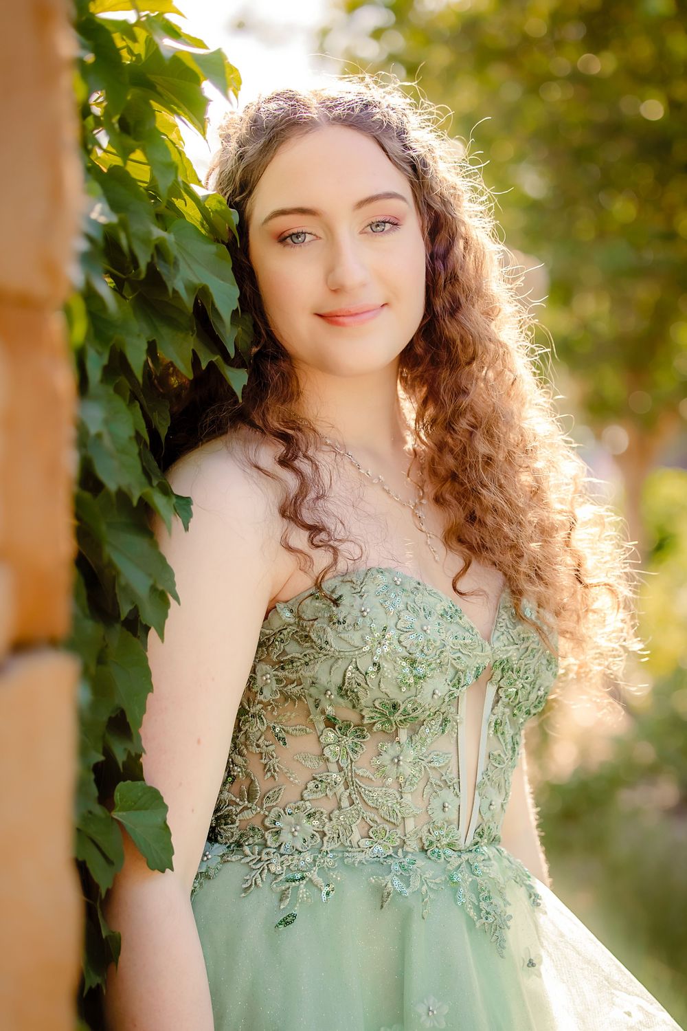 Young woman in green lace gown poses outdoors by ivy-covered wall in golden sunlight with curly hair.