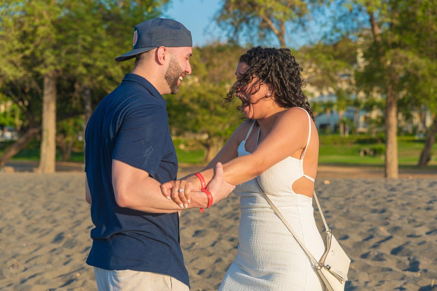 Couple holding hands and smiling at each other on a sandy beach at golden hour.