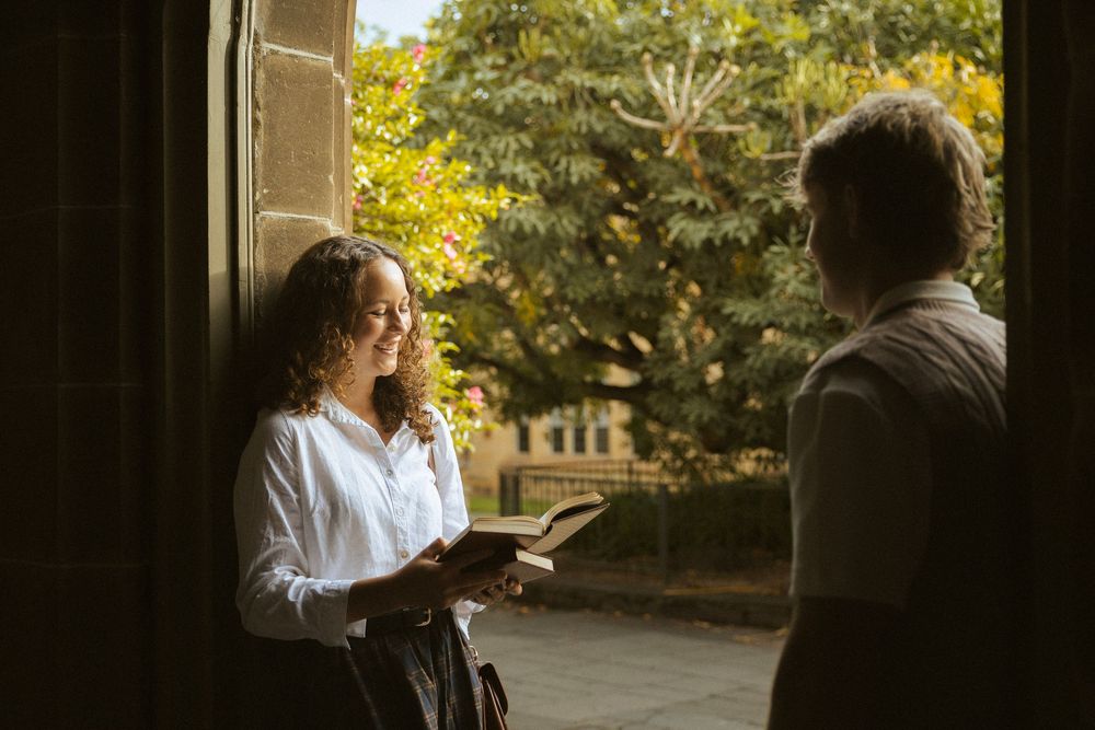 A curly-haired woman in a white blouse holds a book while conversing with a person in a sunlit doorway.