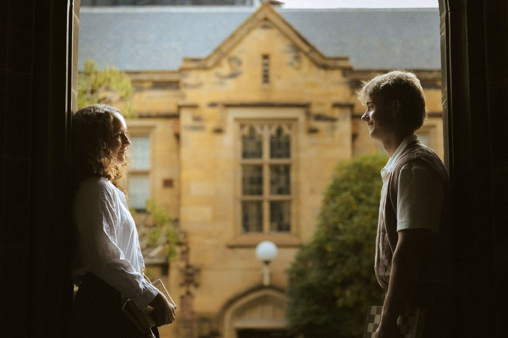 Two people in silhouette conversing by a window overlooking a historic sandstone university building.