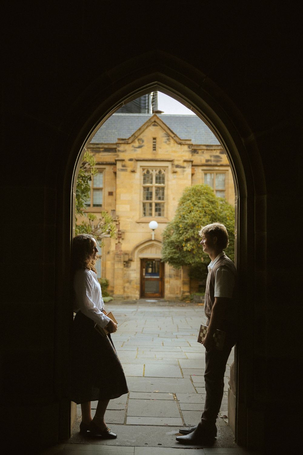 Two students face each other in a Gothic stone archway overlooking a historic university courtyard.