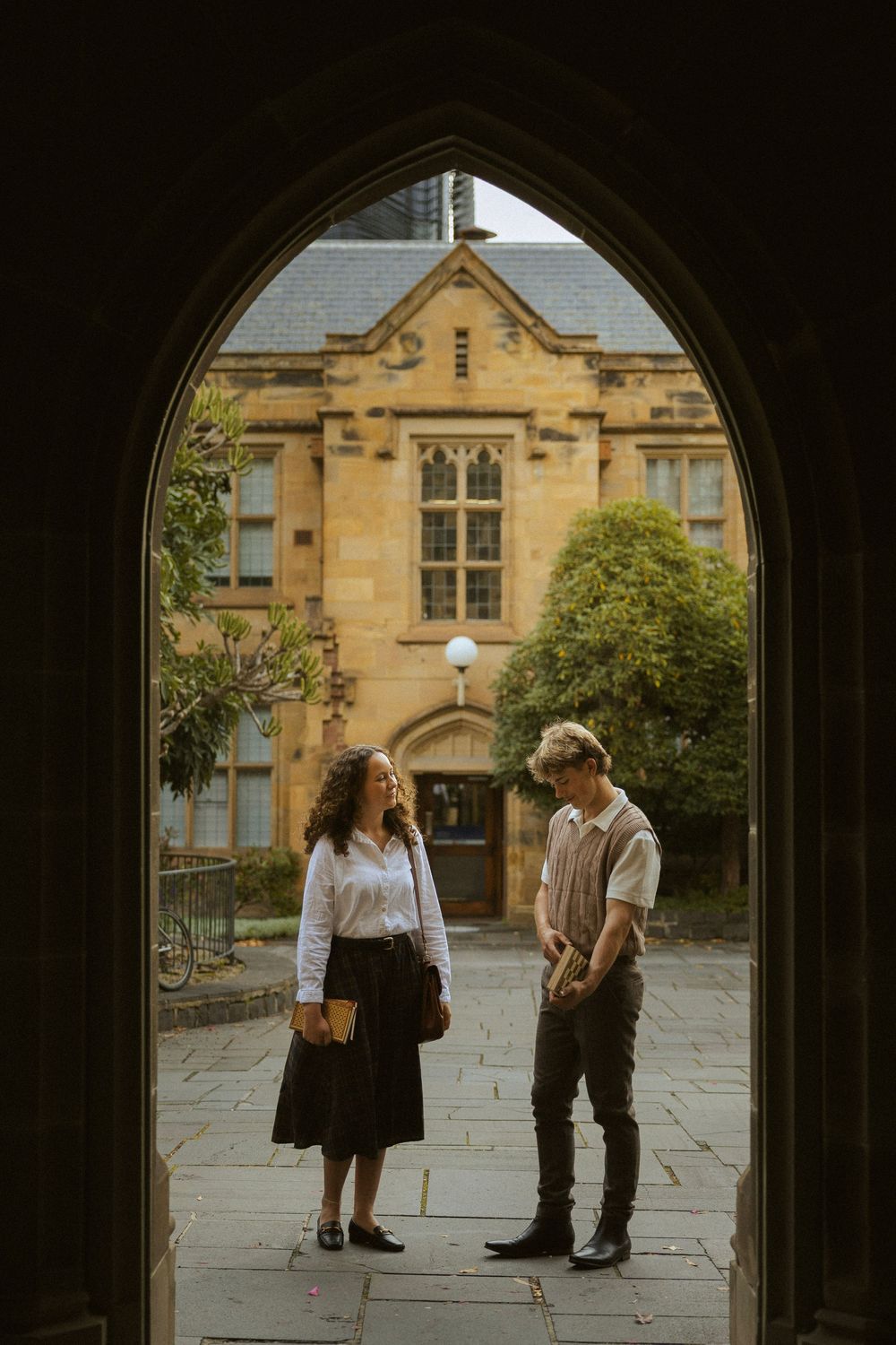 Two students converse in a Gothic stone archway courtyard of a historic university building on a sunny day.