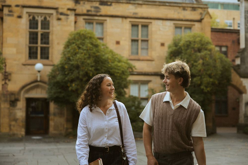 Two students smiling and walking together in a sunlit university courtyard with sandstone buildings.
