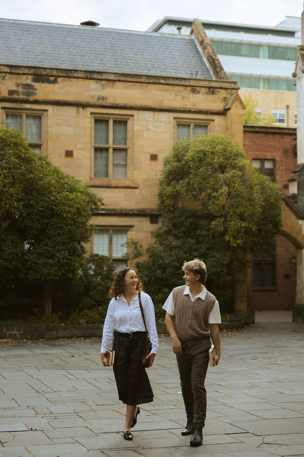 Young man and woman walking and chatting across a stone plaza at a historic university campus.
