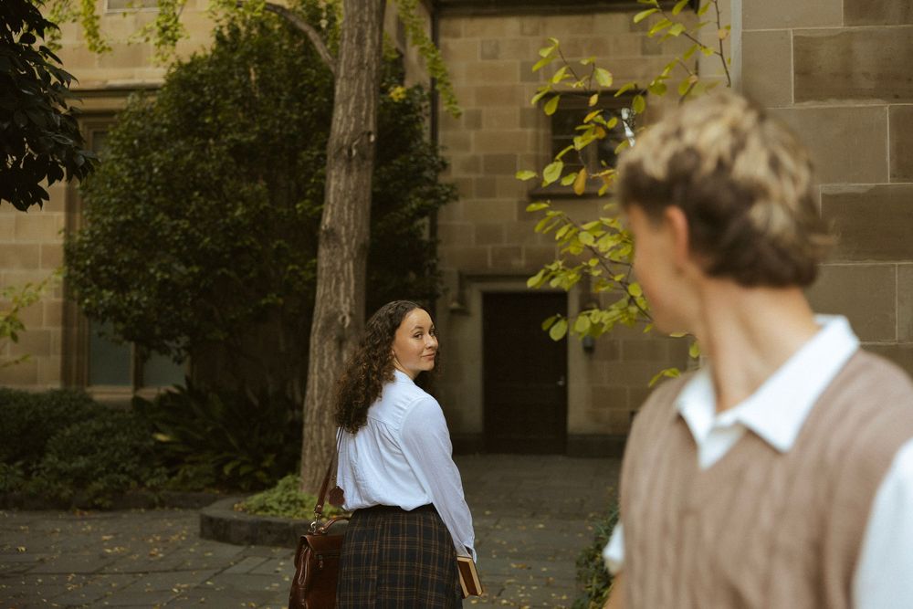 A curly-haired woman leans against a tree glancing back at a man in a courtyard setting.