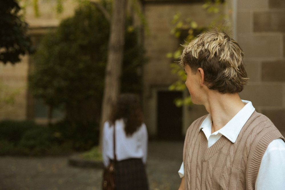 Blonde student in brown knit vest looks across a sunlit university courtyard toward another student walking away.
