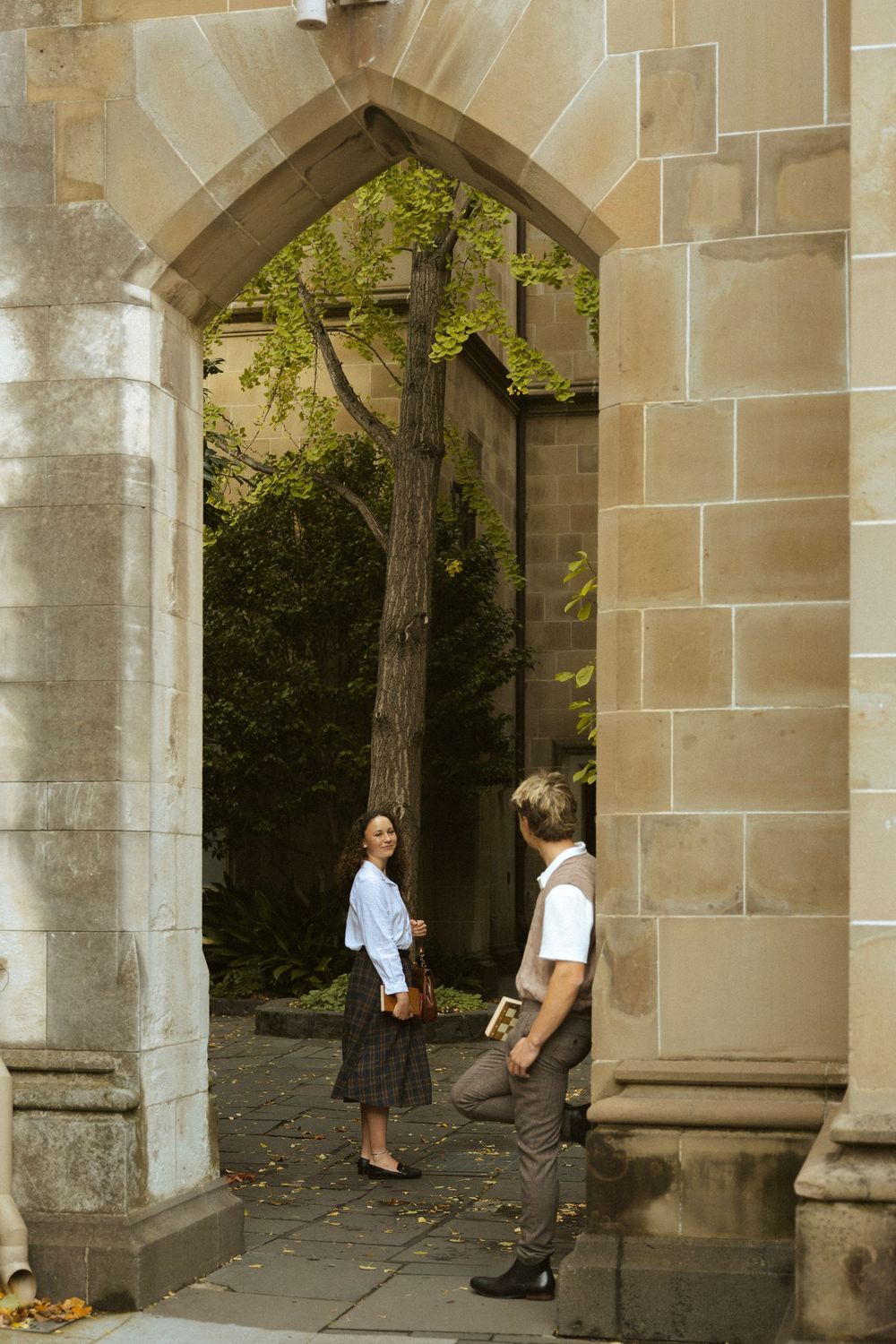 Two students meet under a historic stone archway in a tree-lined university courtyard, one smiling back.
