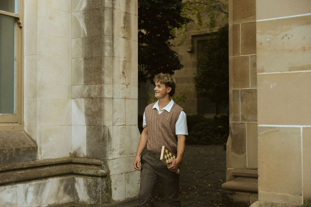 A young man in a brown vest holds a book while leaning against stone columns of a historic university building.