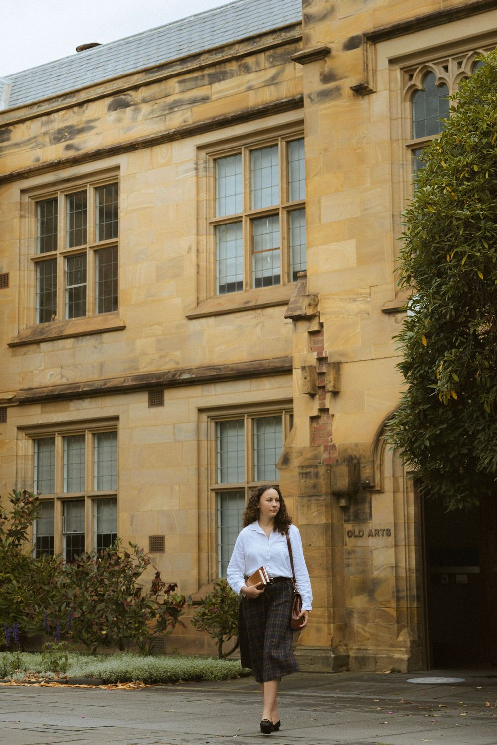 A young woman in a white shirt and dark skirt walks past a historic sandstone university building carrying books.