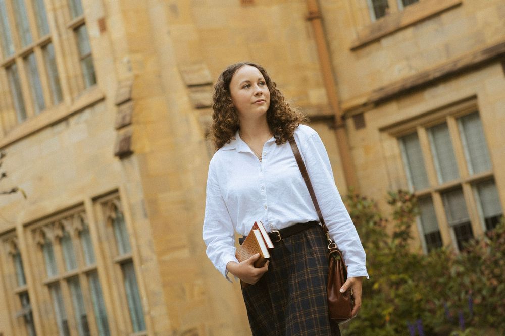 A young woman with curly hair in a white blouse and plaid skirt holds books outside a stone university building.