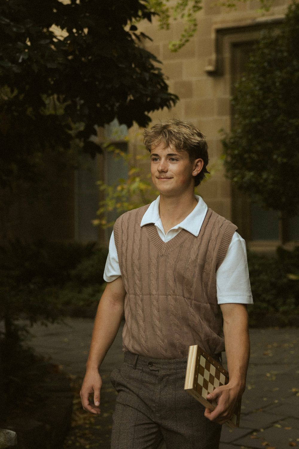 Stylish young male student carrying a book while walking through a lush university campus pathway.