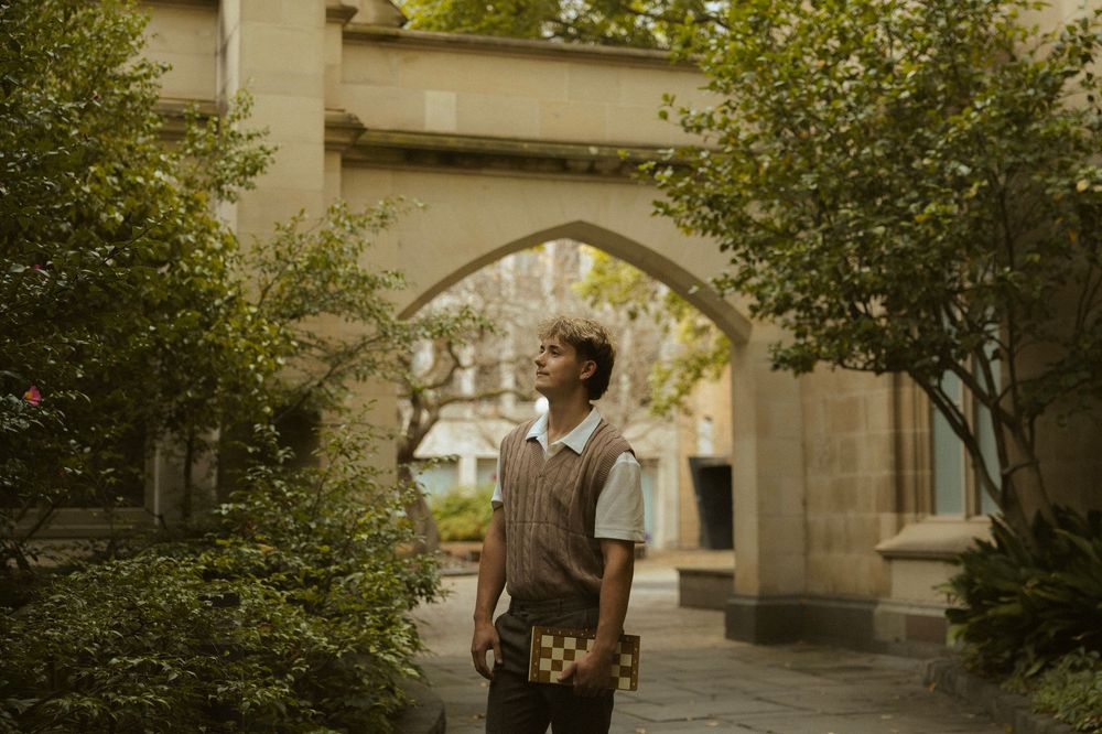 Young man holding a checkered book gazes upward in a lush courtyard with a stone arch.