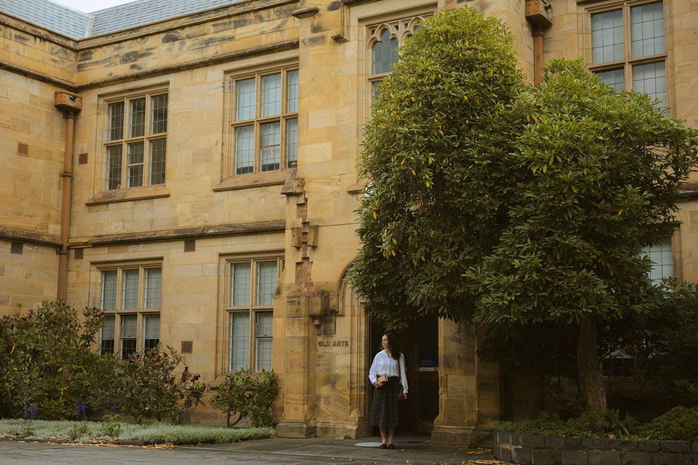 Person standing in arched doorway of historic sandstone university building with lush green trees.