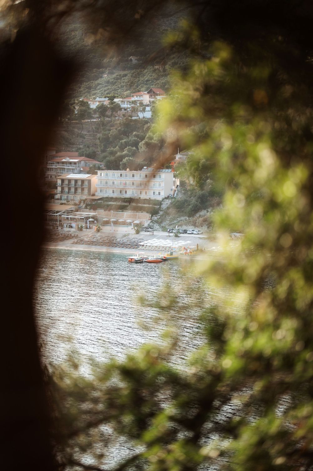 Coastal village with boats on calm water viewed through lush green tree branches in Mediterranean setting.