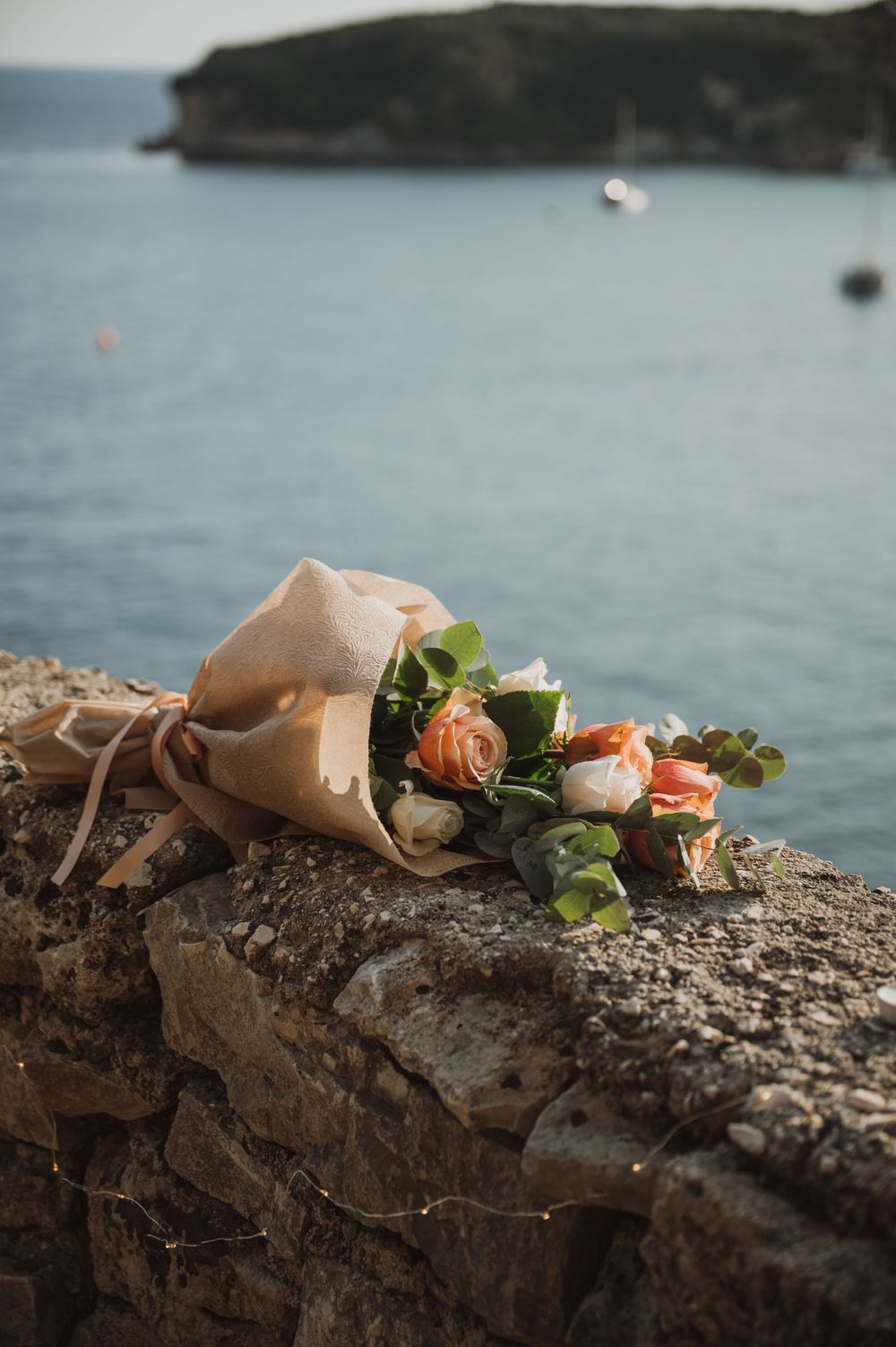 A bouquet of peach roses wrapped in kraft paper rests on a stone wall overlooking a calm blue sea.