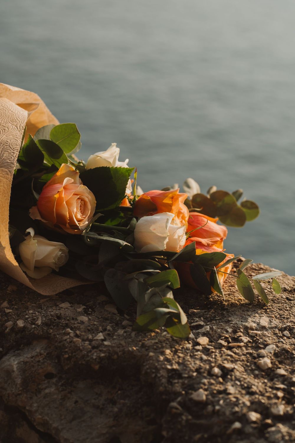 Bouquet of peach and white roses wrapped in kraft paper resting on rocky seaside cliff.