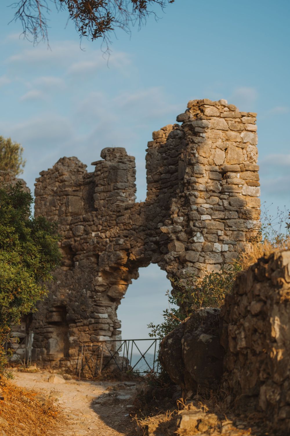 Ancient stone ruins with arched gateway surrounded by trees under blue sky in Mediterranean.