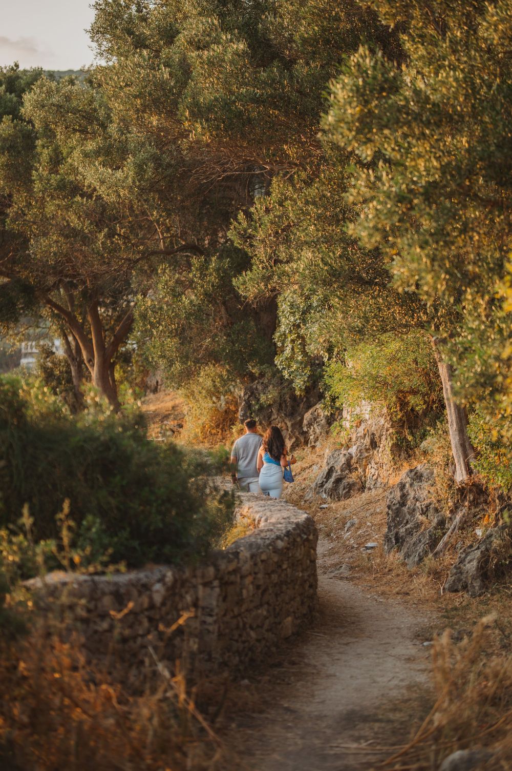 Couple sitting on a stone wall path surrounded by olive trees at golden hour in a Mediterranean landscape.
