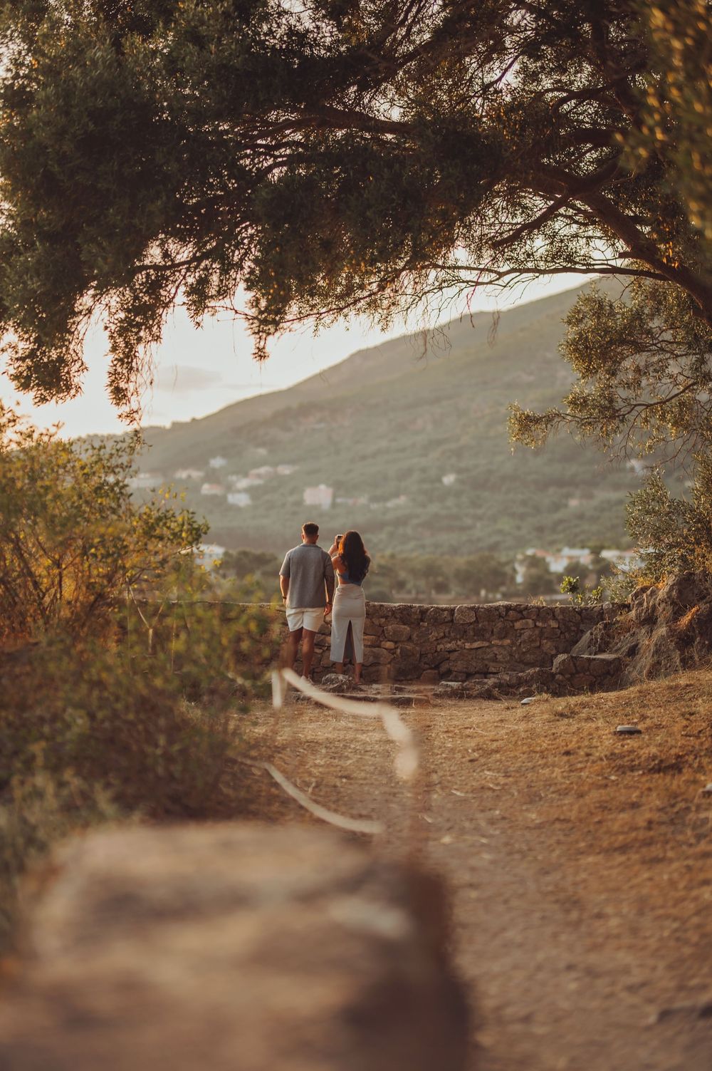 Couple walking hand in hand on a scenic hilltop path surrounded by trees at golden hour.