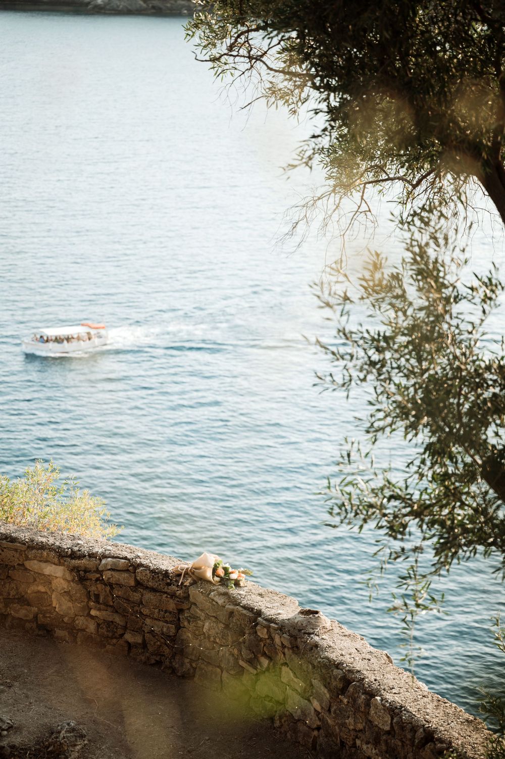 A scenic coastal view with a boat on blue water, stone wall, dog resting, and olive tree branches in sunlight.