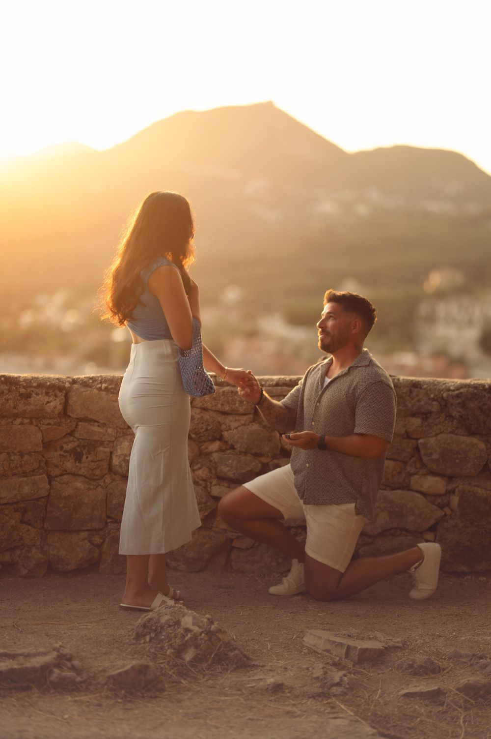 Man on one knee proposing to woman at sunset with mountain village backdrop.