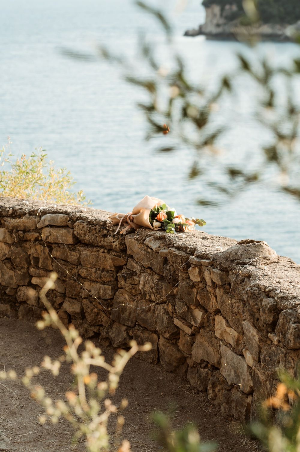 Bouquet of flowers resting on ancient stone wall overlooking serene blue sea at golden hour.