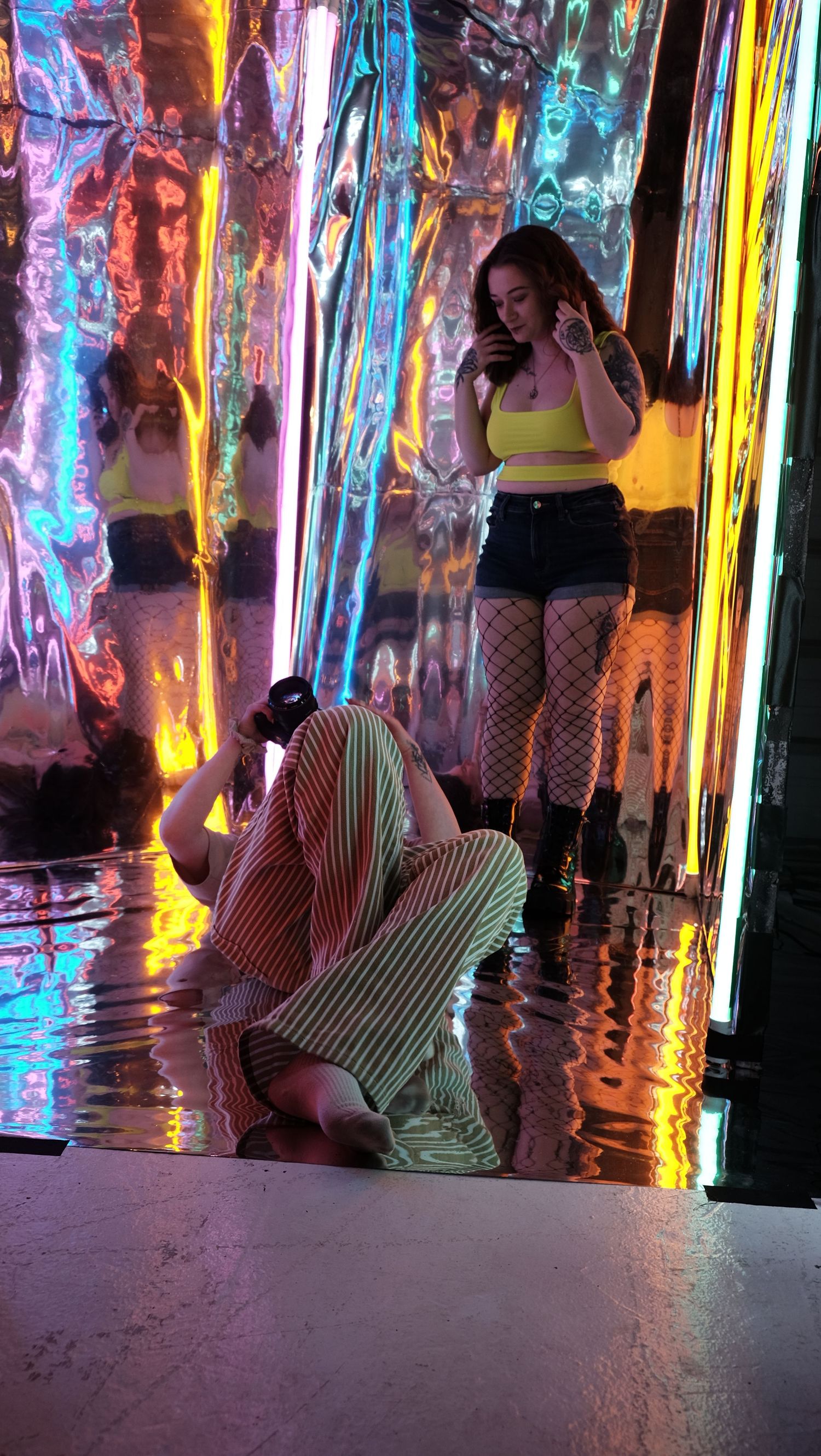 Two women pose in a vibrant holographic installation with colorful reflective curtains and neon lighting.