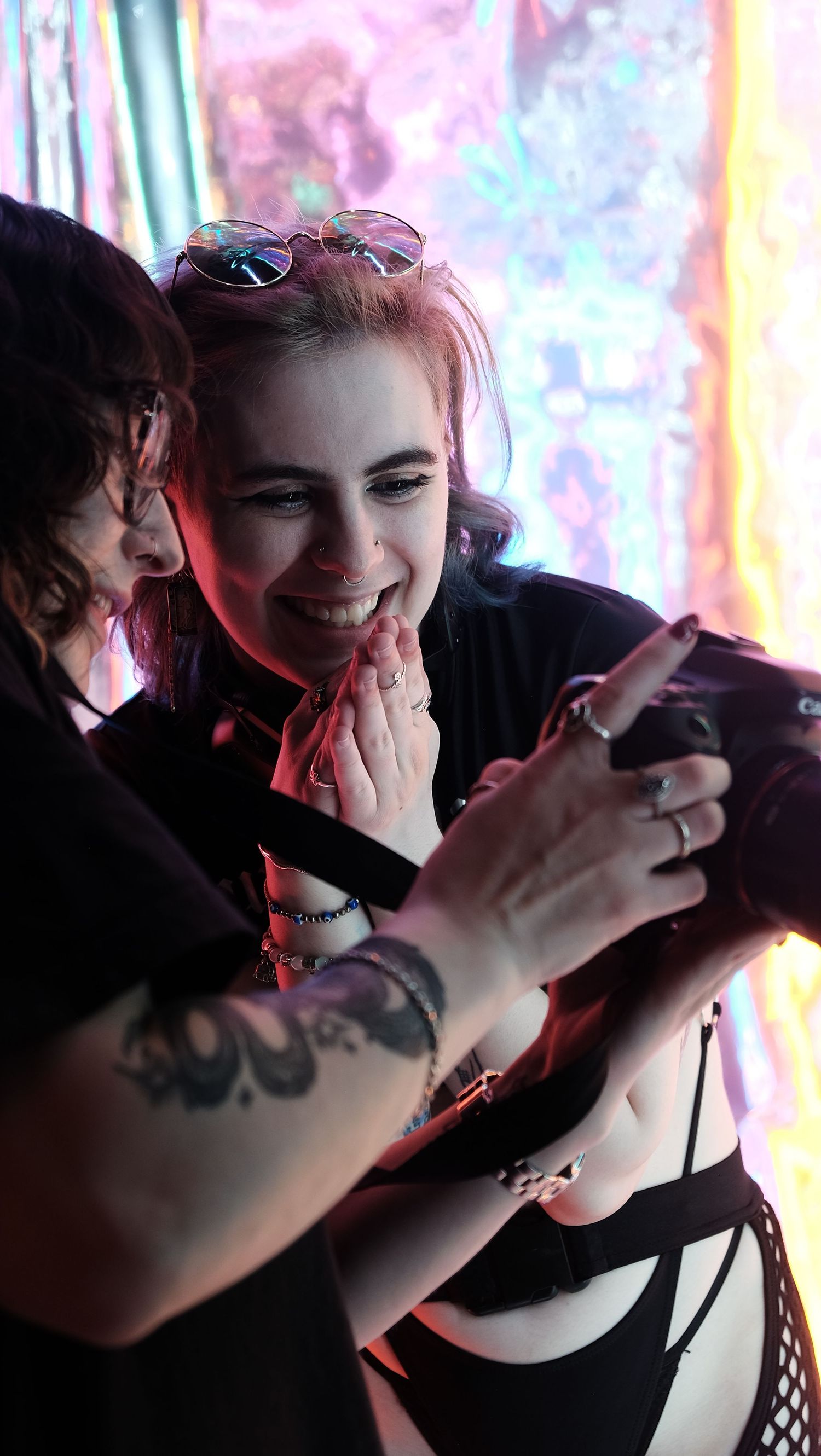 Two women with tattoos and edgy style laugh together while looking at a camera at a colorful event.