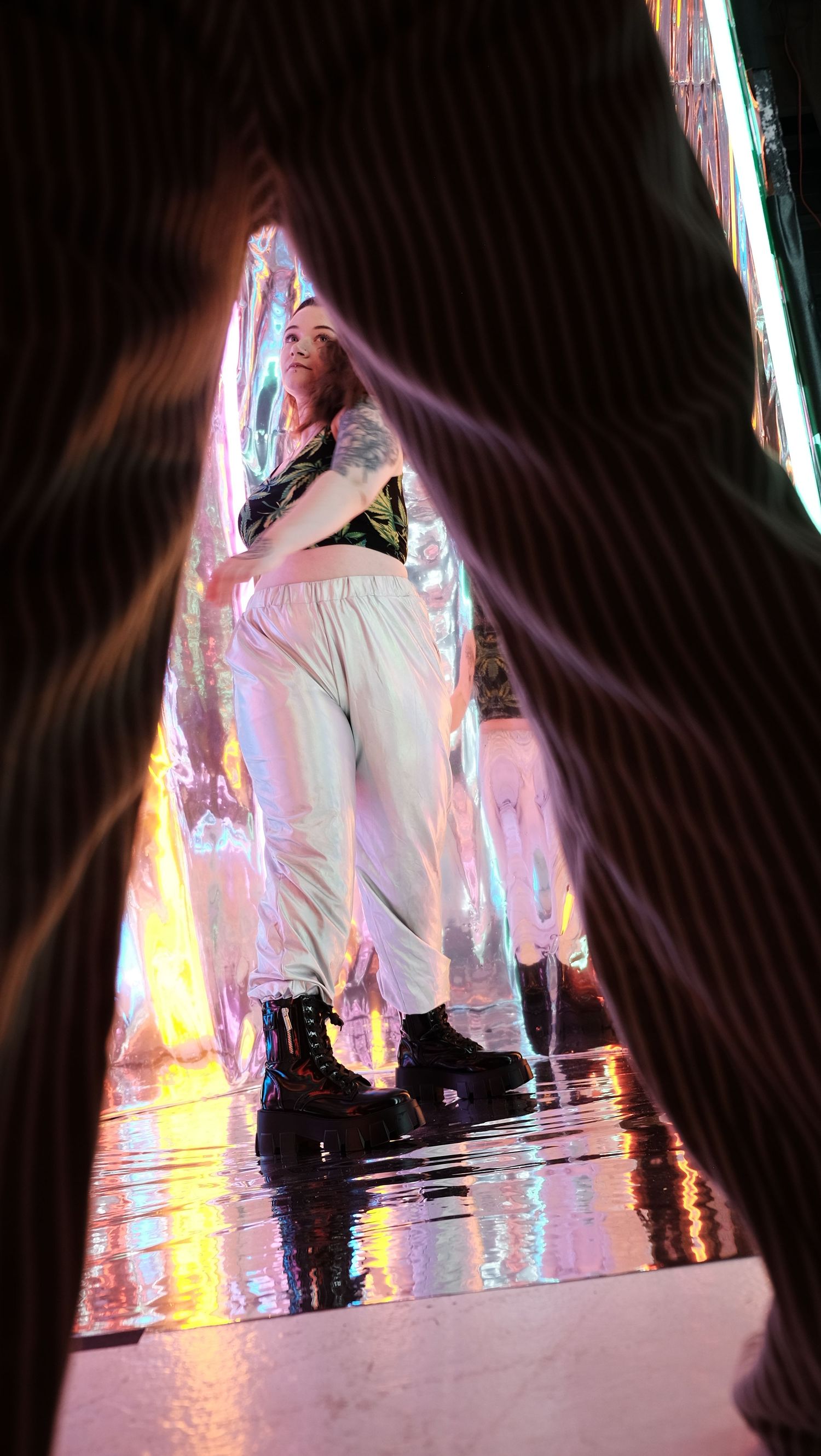 Woman in white pants and black boots stands in colorful iridescent light installation, viewed through a dramatic framing gap.