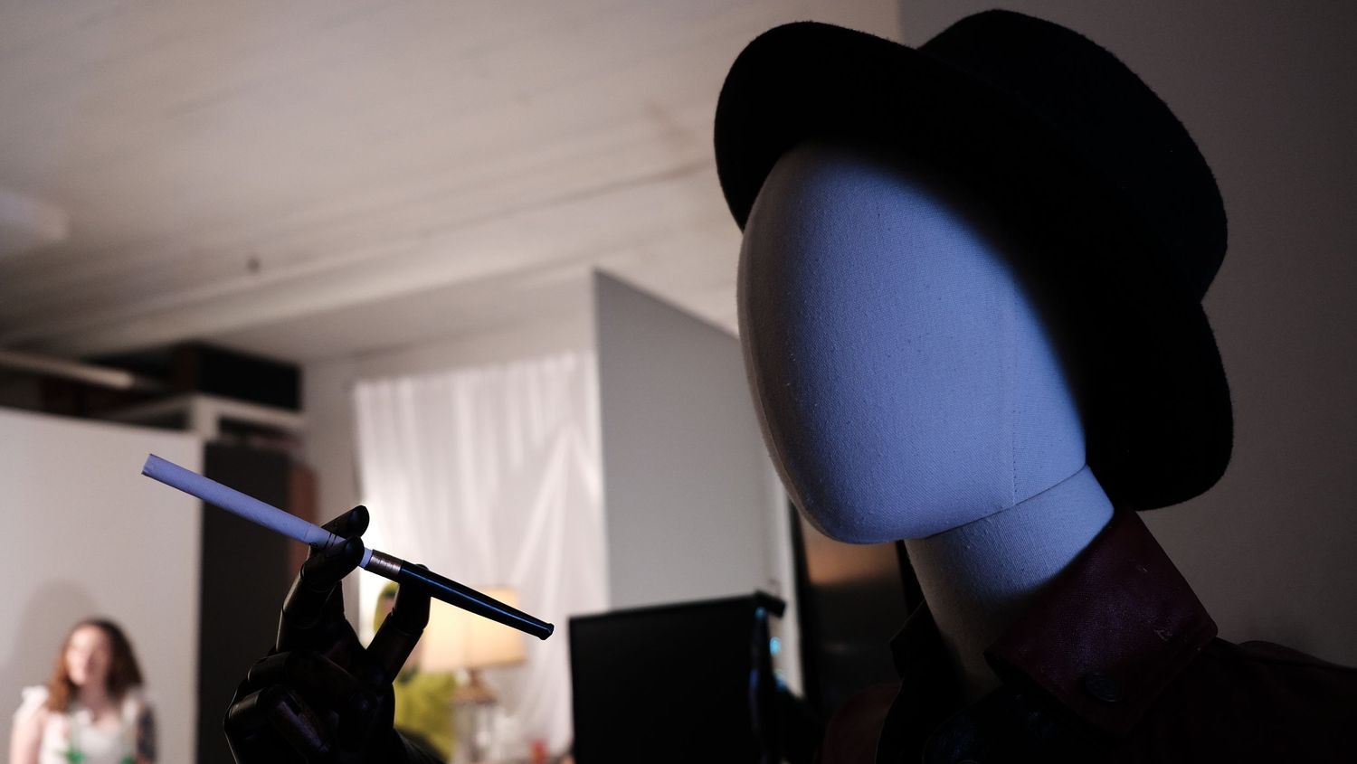 A mannequin wearing a black bowler hat and glove holds a pen in a dimly lit room with a person in the background.