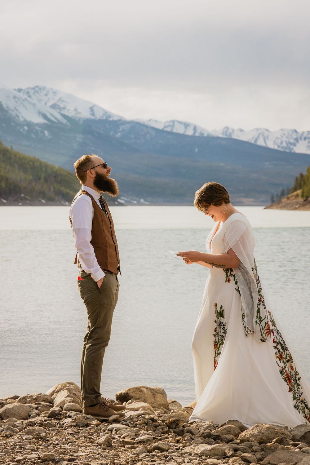 Bride reads vows to bearded groom by a rocky lakeside with snow-capped mountains backdrop at outdoor wedding.