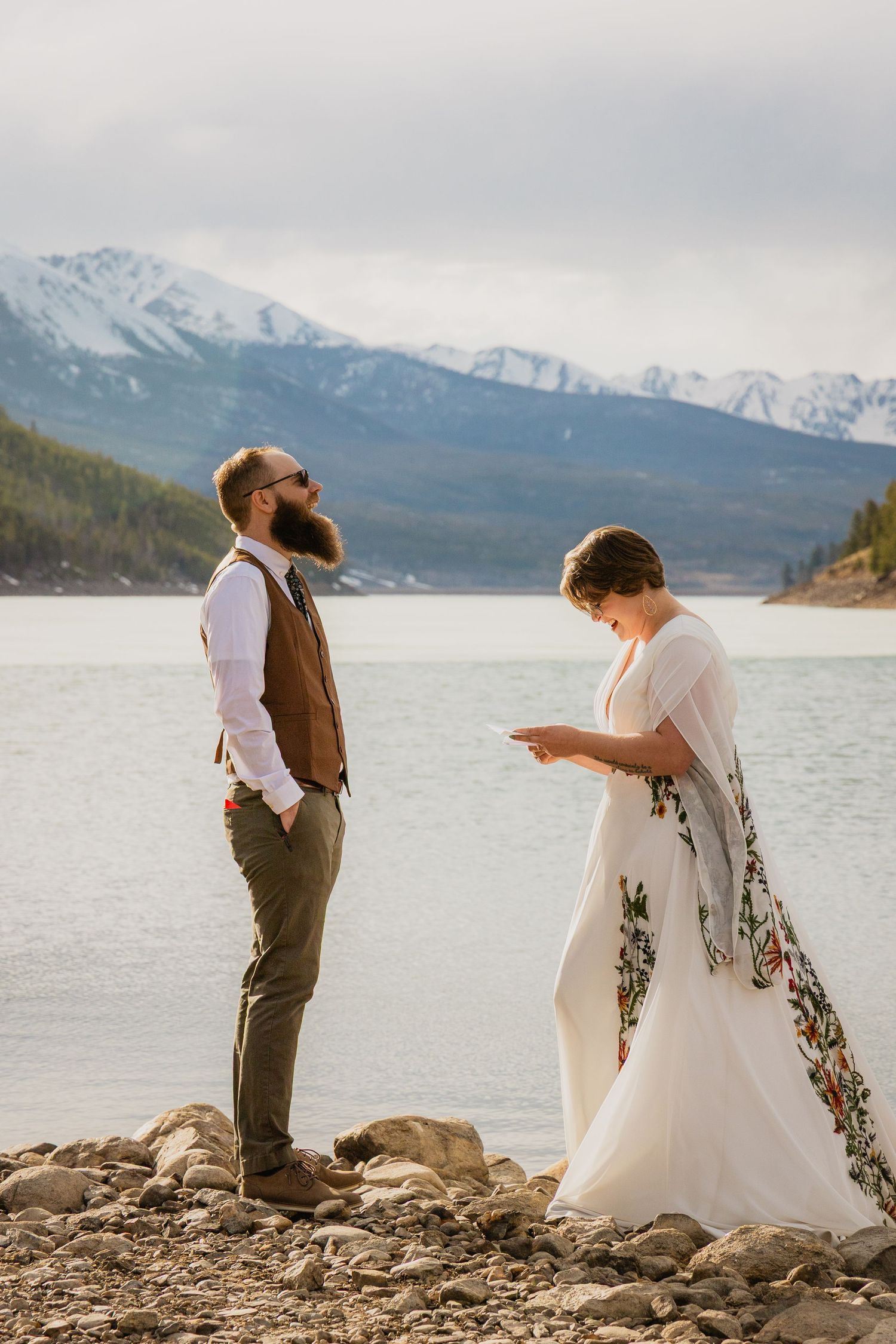 Bride reads vows to bearded groom by a rocky lakeside with snow-capped mountains backdrop at outdoor wedding.